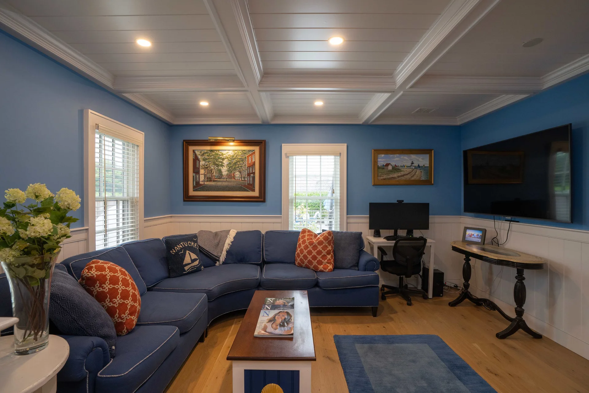 The Captain’s Quarters family room: a curved navy sofa with rust and Nantucket-motif pillows under a white coffered ceiling, blue walls hung with framed Nantucket street and beach paintings, a wall-mounted TV, and a small desk with a monitor in the corner.