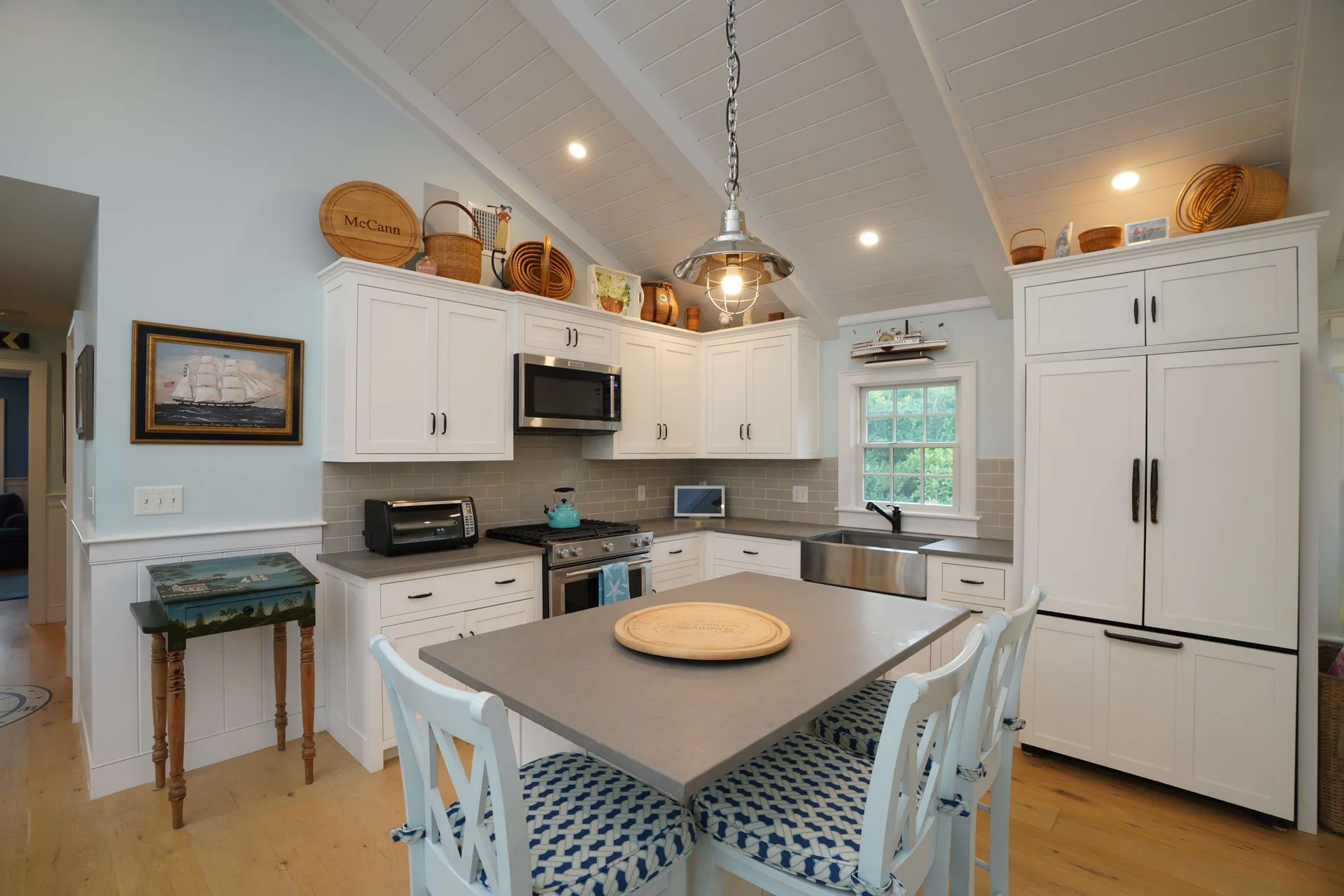 The Captain’s Quarters kitchen: white shaker cabinets under a vaulted white shiplap ceiling, gray subway tile and stone counters, a stainless farmhouse sink, lightship baskets along the cabinet tops, and a stone-topped island with cushioned chairs beneath a pendant light.