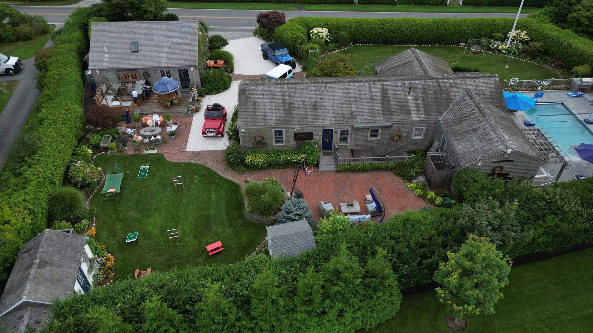 An oblique aerial of the Mini Compound: the Captain’s Quarters cottage with its outdoor-kitchen deck and blue umbrella, the brick fire-pit lounge, vintage Jeeps on the drive, a games lawn with a hammock and Chippo, and the pool just visible at the edge.