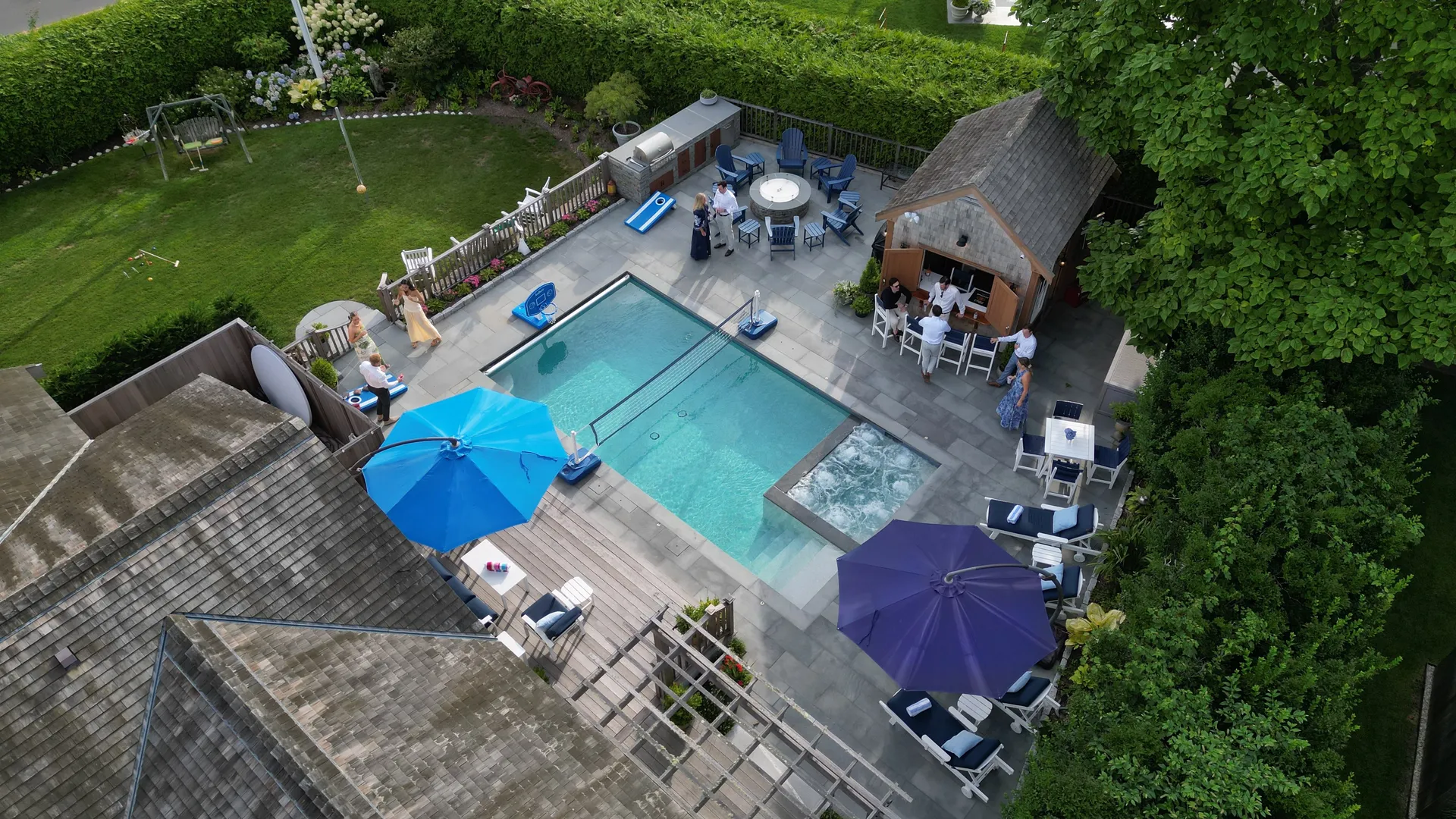 Another aerial of the pool and spa with the volleyball net strung across it, the cabana bar with guests on stools, the outdoor kitchen and grill, an American flag on the shed, and croquet set up on the adjoining lawn.
