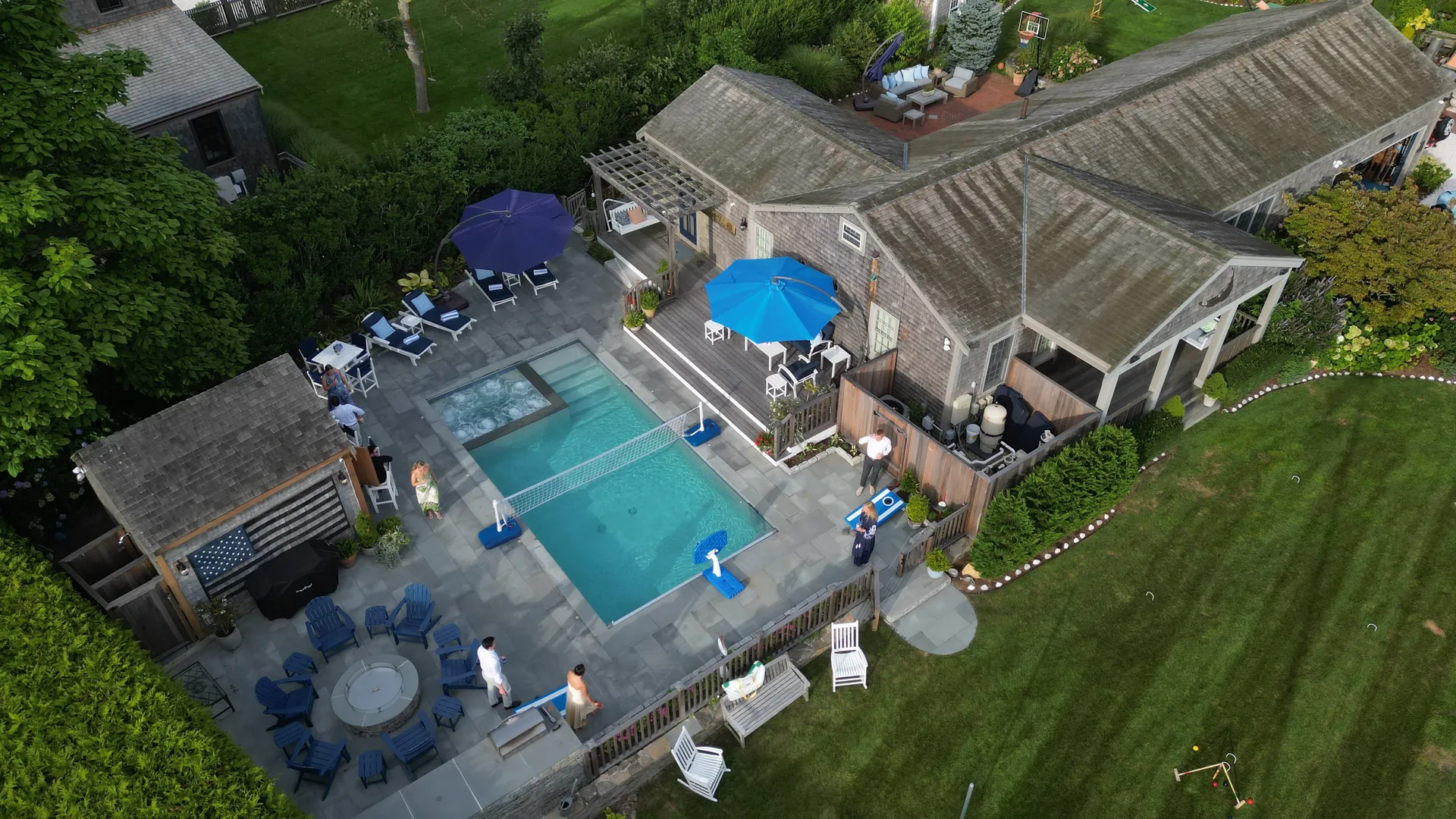 Aerial view of the private Mini Compound pool: a bluestone-wrapped pool with a built-in spa and a volleyball net across the water, blue and purple umbrellas over navy loungers, a shingled cabana bar and an outdoor kitchen along one side, and a fire-pit ring of navy Adirondack chairs on the terrace.