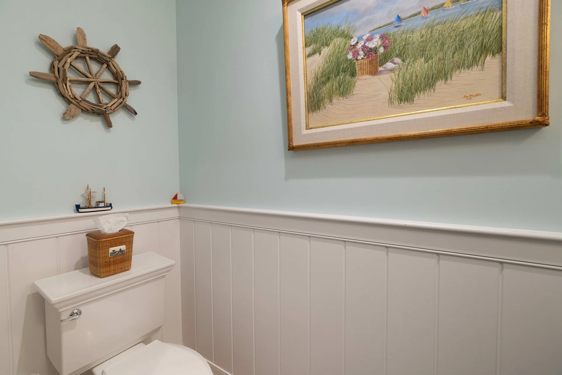 Another corner of the same powder room: a ship’s wheel made of weathered driftwood on the sea-glass wall, small model sailboats lined along the chair rail, and the framed catboats pastel alongside.