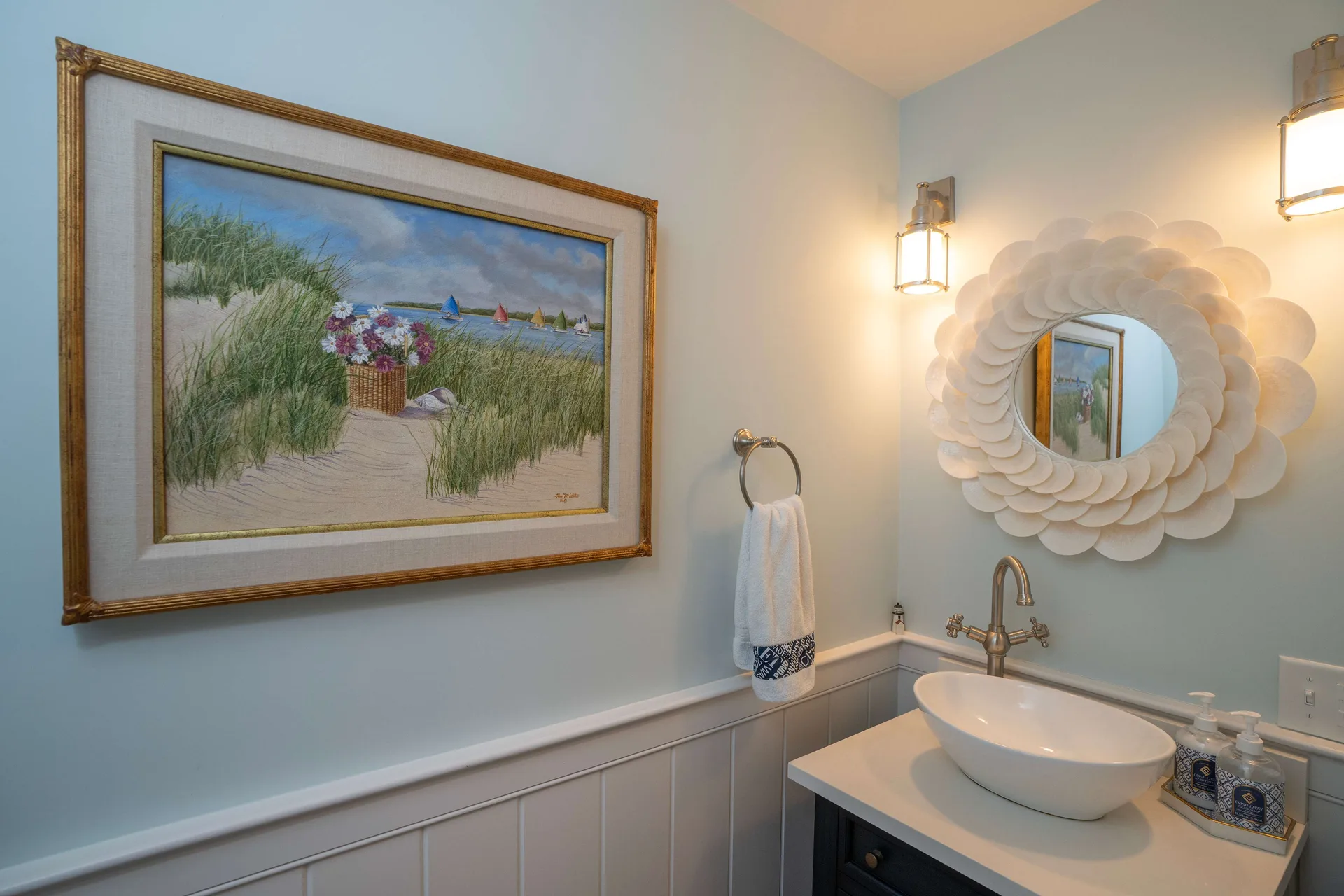 The Captain’s Quarters powder room: sea-glass walls with white beadboard wainscot, a round mirror ringed with overlapping capiz shells between two nautical sconces, a white vessel sink on a navy vanity, and a gold-framed pastel of dune grass and catboats on the wall.