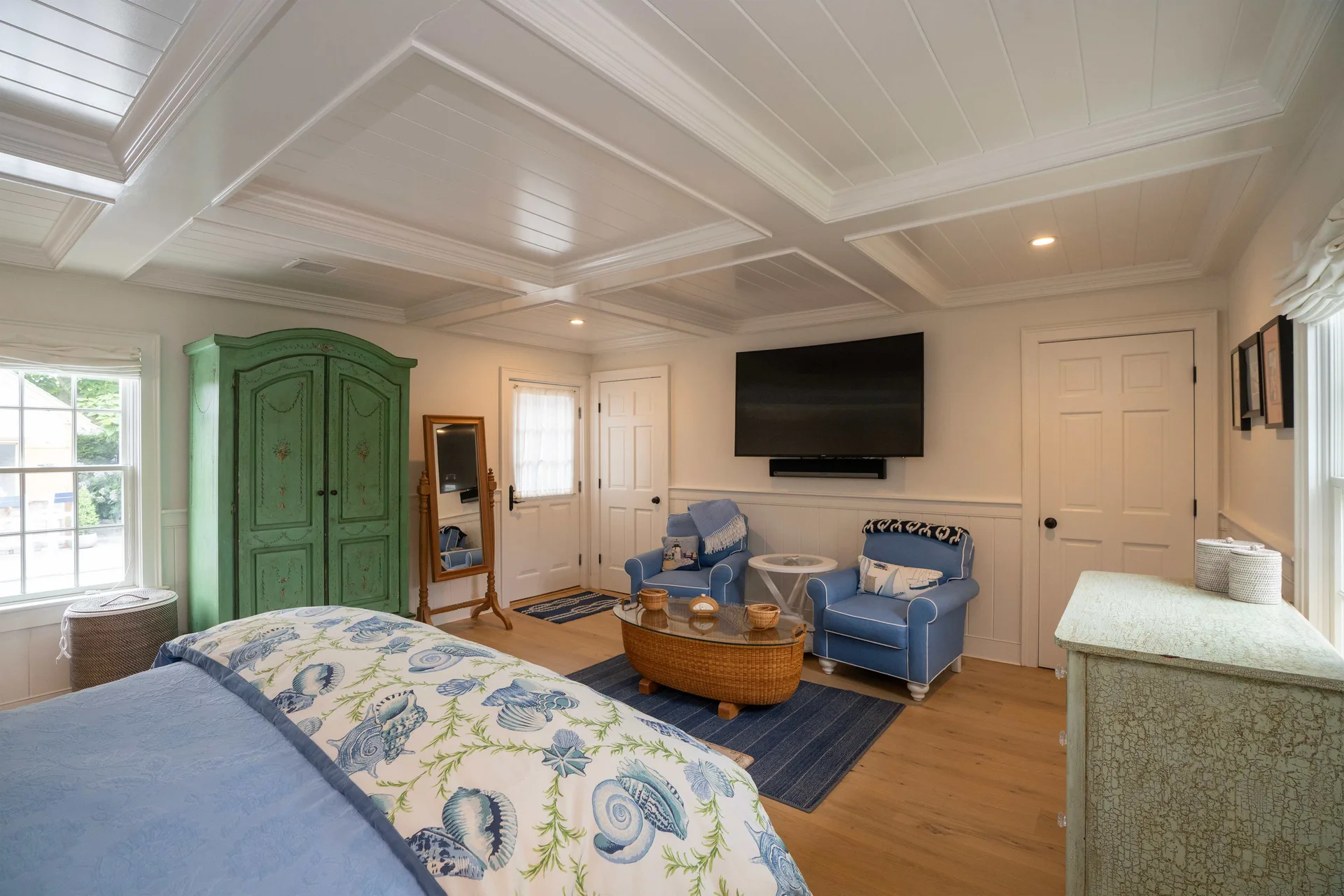 The sitting area within the same suite: two blue upholstered armchairs around a round wicker coffee table, with the green armoire and cheval mirror beyond.