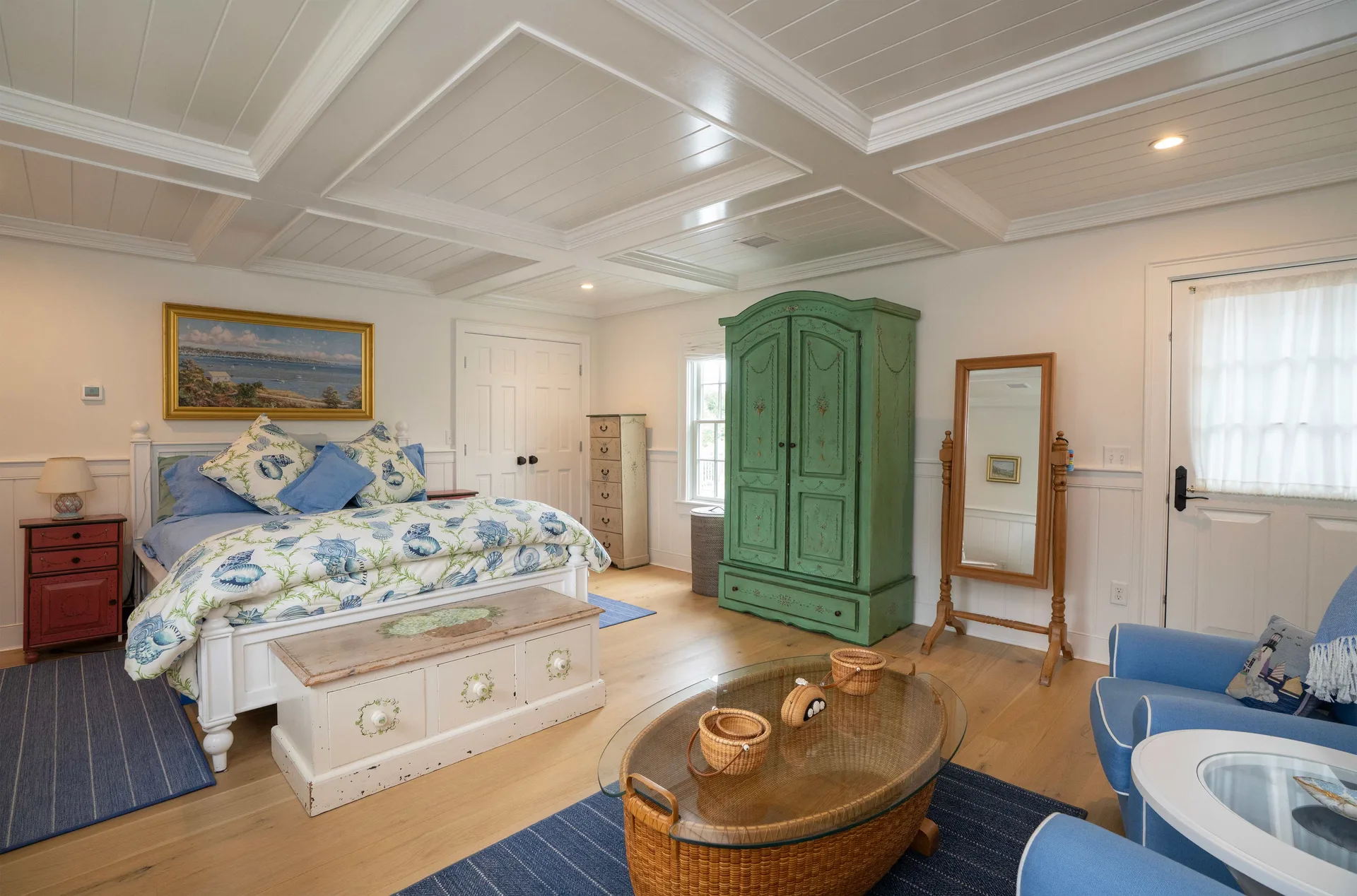 The Captain’s Quarters primary suite: a white four-poster bed in a blue-and-green seashell duvet under a coffered ceiling, a hand-painted green armoire, a cheval mirror, a red painted nightstand and a painted blanket chest at the foot of the bed.