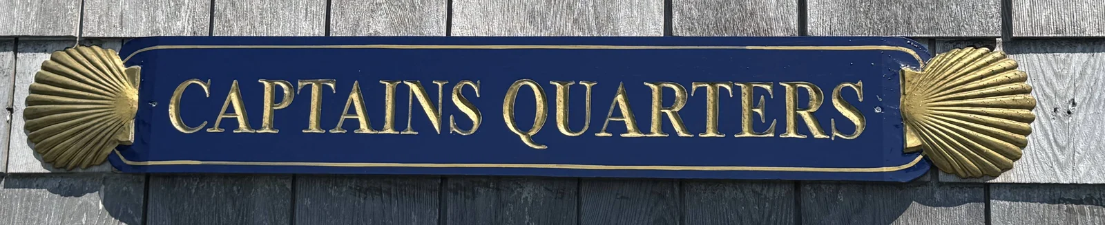 The Captain’s Quarters house sign: a navy carved quarterboard with “CAPTAINS QUARTERS” in gold gilt capitals, flanked by a gold carved scallop shell at each end, mounted on weathered gray cedar shingles.