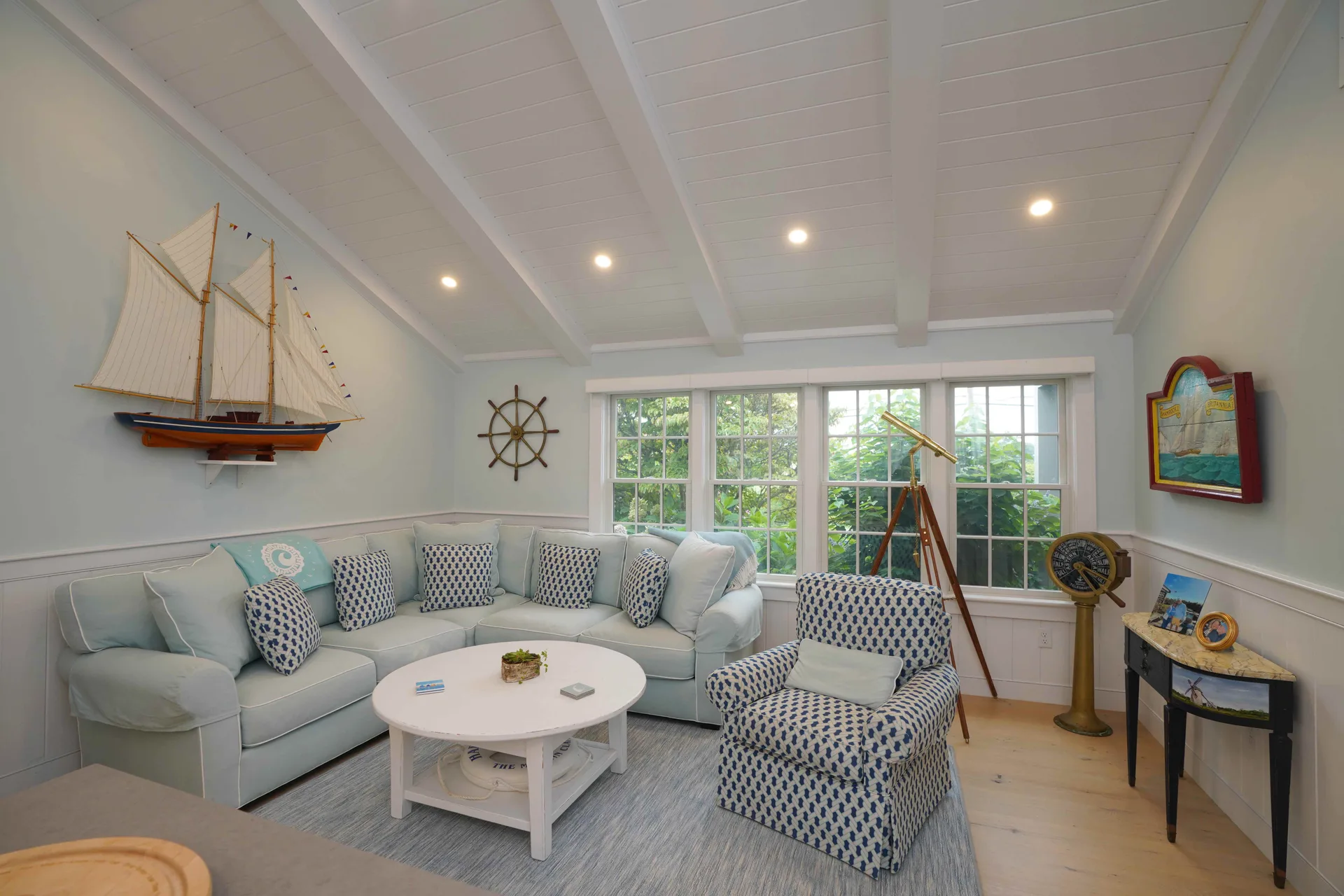 Captain’s Quarters: the open sitting room off the kitchen under a vaulted white shiplap ceiling — a pale-blue sectional and armchair, a model sailboat, a ship’s wheel and a brass telescope at the windows, with the kitchen island edge in the foreground.