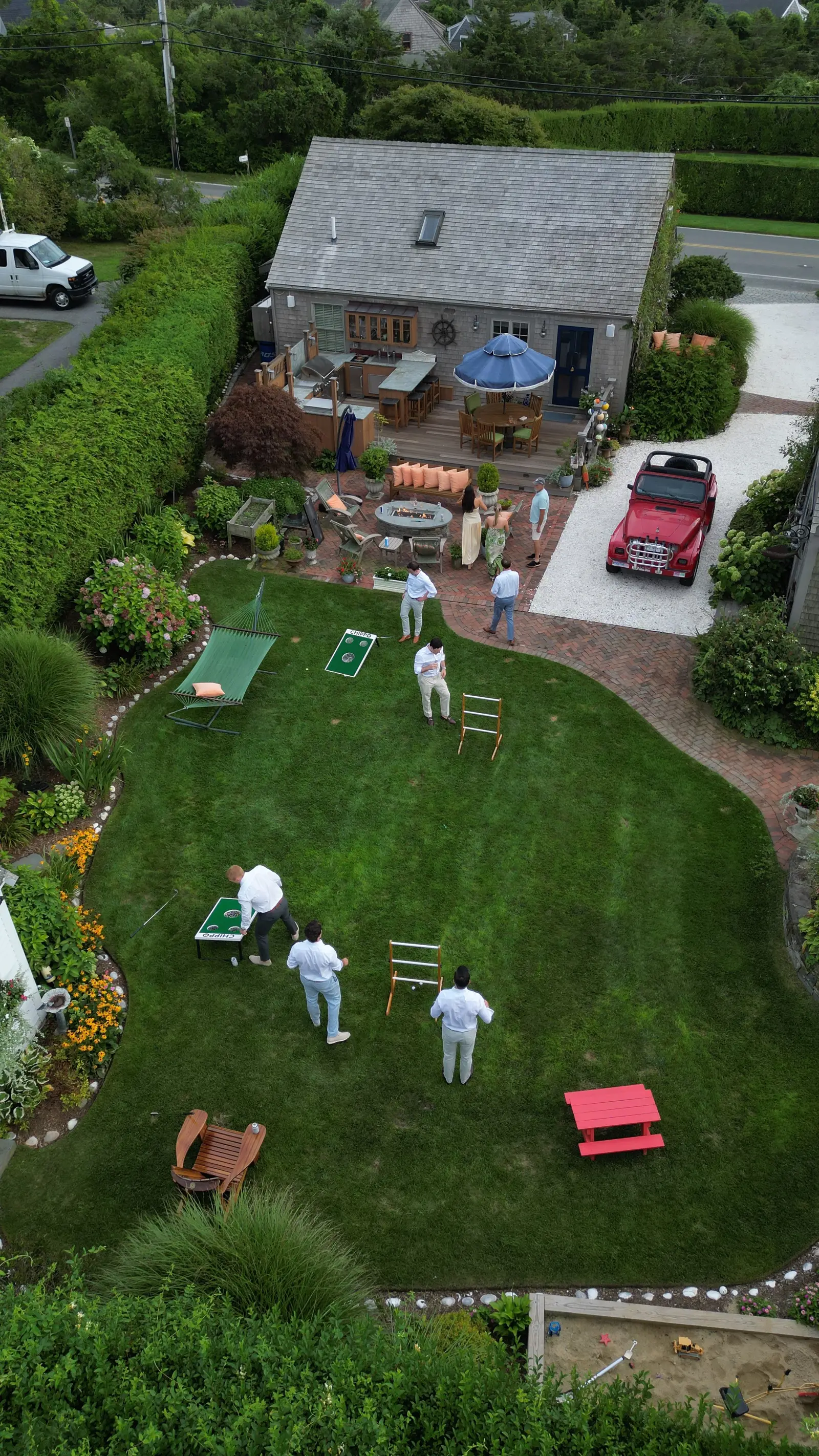 Another aerial of the Crew yard showing the deck outdoor kitchen and blue-umbrella dining, the fire-pit lounge with orange cushions, a red vintage Jeep on the drive, and the lawn dotted with cornhole, ladder toss and the hammock.