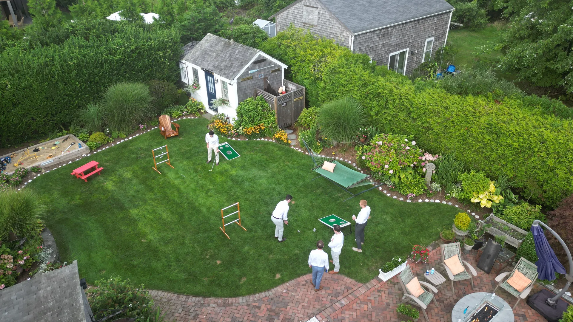 Aerial view of the back lawn set up for games: a Chippo golf board and ladder-toss frames on the grass, a green hammock, a red kids’ picnic table, Jack’s sandbox in the corner, and the brick fire-pit patio by the cottage, with guests playing across the yard.