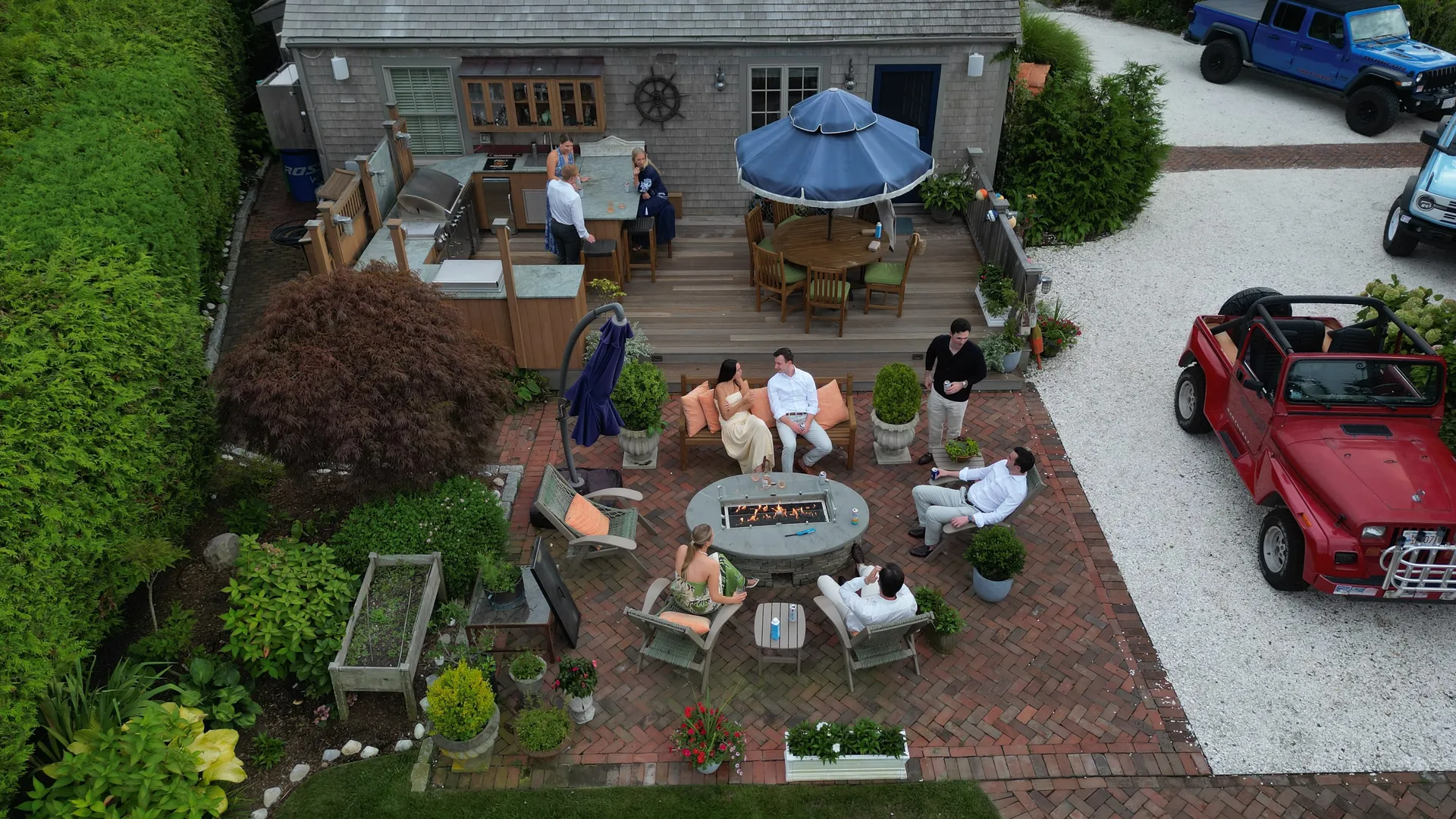 Aerial view of the herringbone brick fire-pit terrace: the round stone gas fire table lit at the centre, ringed by orange-cushioned wicker chairs and a bench, with the outdoor kitchen and bar on the deck behind and vintage Jeeps parked on the drive alongside.