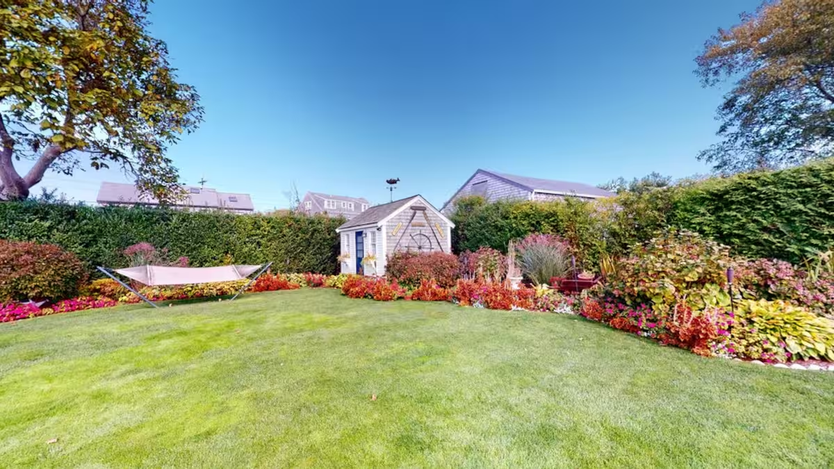 The back lawn at Crew with a hammock strung between trees, a small shingled garden shed topped by a whale weathervane, and deep flower borders along the grass.