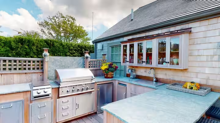 Built-in outdoor kitchen set into a shingled wall: stainless grill and side burner, refrigerator drawers, a sink in pale stone counters, glass-front upper cabinets and teal-painted shutters.
