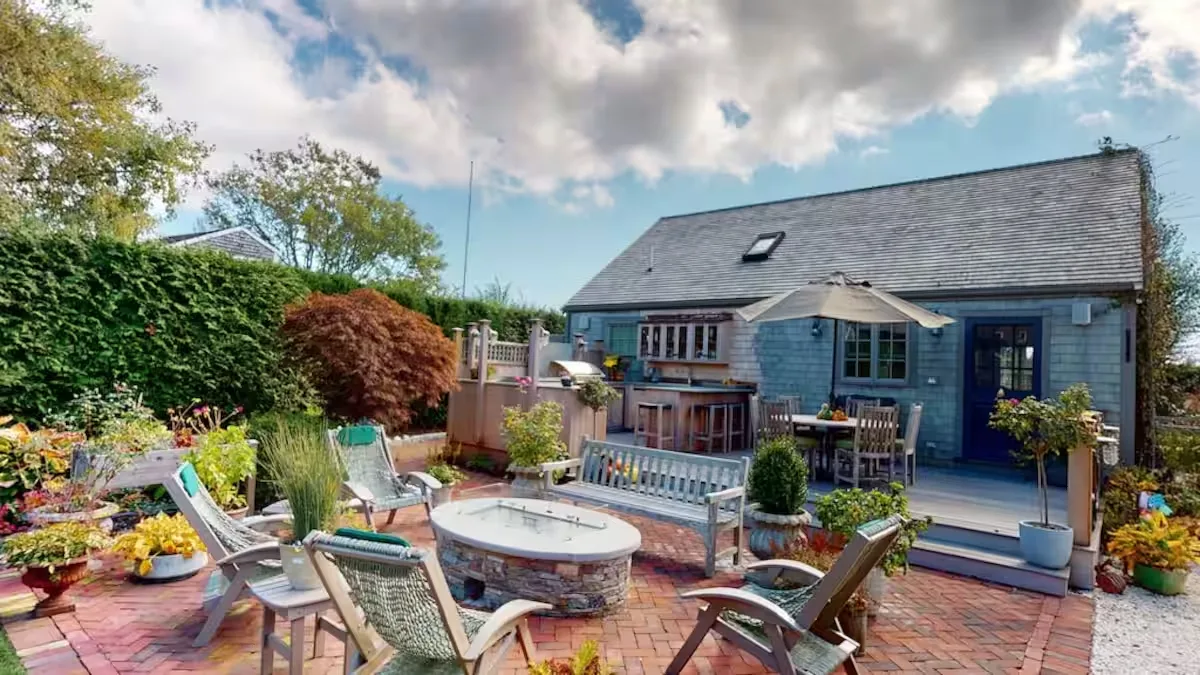 Crew at Nantucket: the cedar-shingled cottage rising behind a brick patio with a firepit lounge and outdoor kitchen, under a dramatic cloud-swept sky.