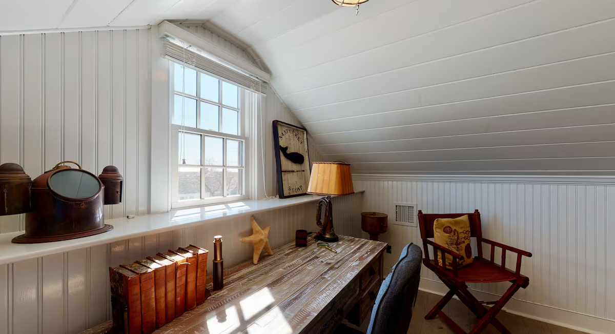 The desk nook under the eaves: a reclaimed wood table below a dormer window, an antique brass ship’s binnacle and a row of leather-bound books on the sill, a Nantucket Sound whale sign, and a red wooden director’s chair