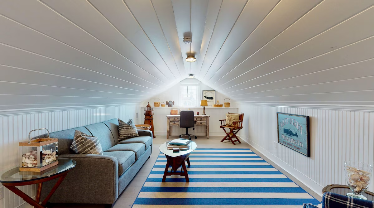 The top-floor sitting room under a barrel-vaulted white plank ceiling, with beadboard walls, a blue-gray sofa, a blue-and-white striped rug, an oval coffee table stacked with books, and a reclaimed wood desk in the dormer window at the far end beneath a Nantucket Whaling Co sign