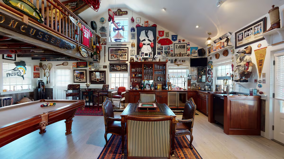 The bar end of the barn: a mahogany bar with beer taps and a mirrored back bar of bottles, a red barber’s chair, a kegerator and undercounter fridges, a boxing display and Yankee Stadium sign above, and the pool table in the foreground