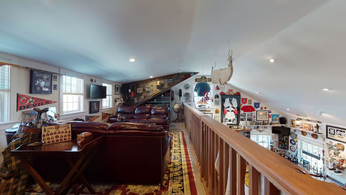 The barn loft: a maroon leather sectional and wall-mounted television under the eaves, a New Jersey Devils pennant, golf clubs and framed photographs, a carved white goose hanging from the rafters, and the balcony rail looking down over the bar