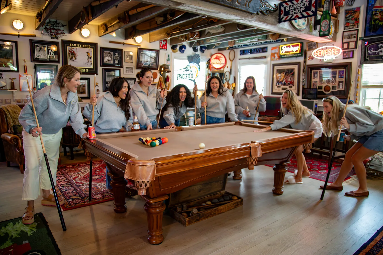 A group of friends in matching gray quarter-zips laughing around the barn’s slate pool table mid-game, cues in hand, under the memorabilia-covered walls and neon beer signs with oars mounted across the ceiling beams