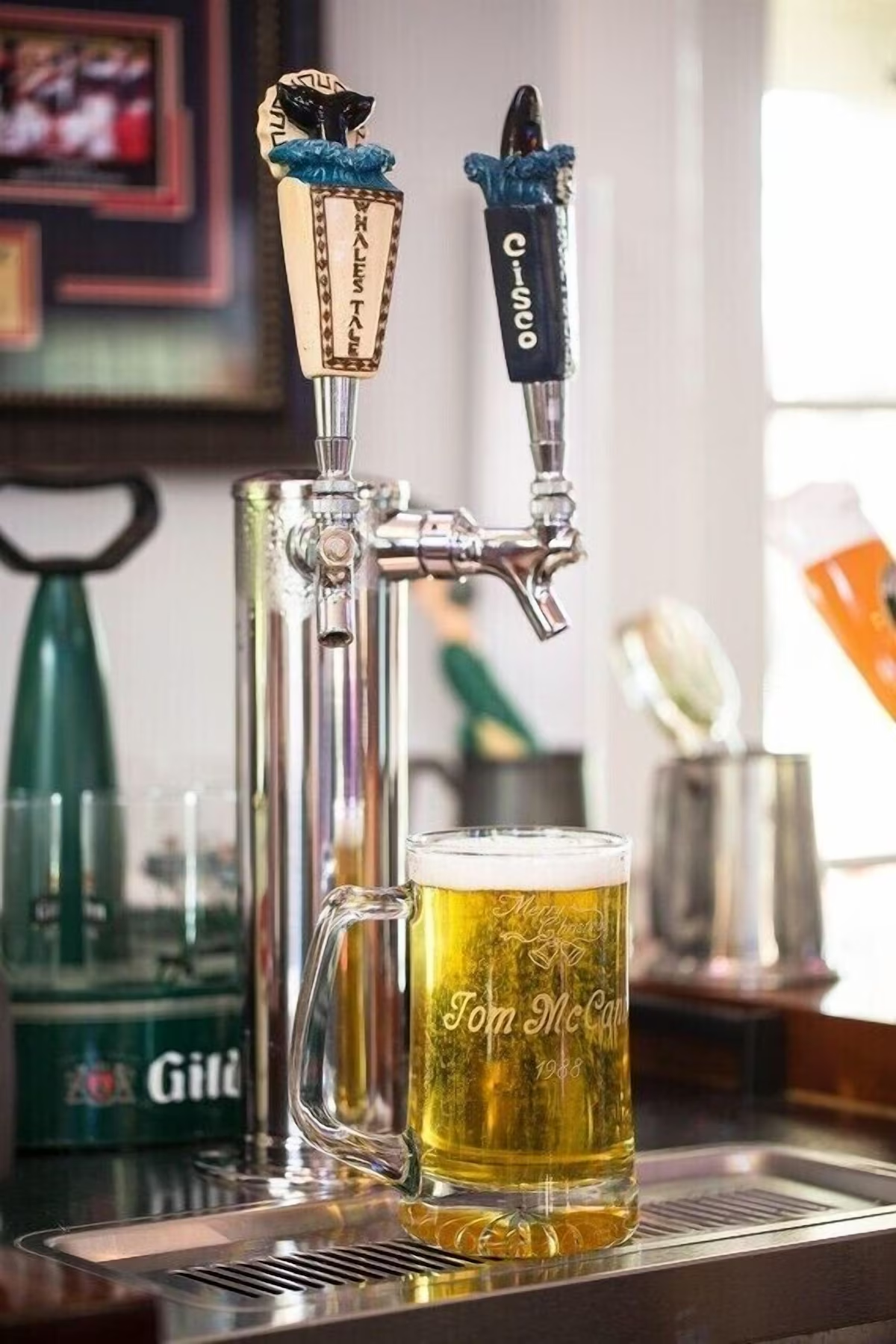 Two tap handles at the barn bar — a scrimshaw-style Whale’s Tale and a black Cisco, both collared in blue glaze — over a full glass mug engraved “Tom McCann 1988” on the drip tray