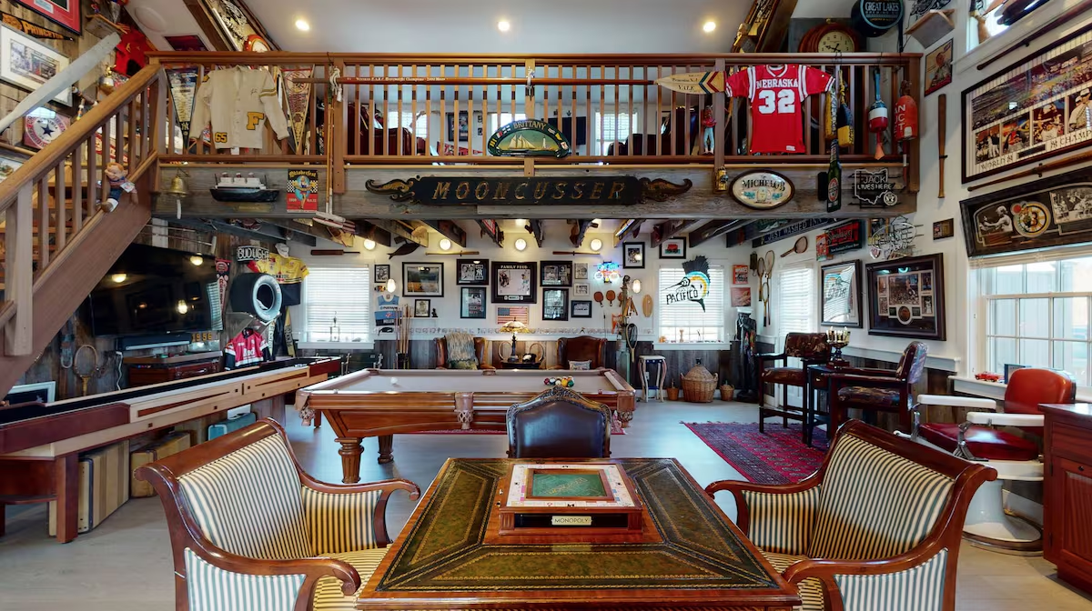 The barn from the card table: a slate pool table and shuffleboard on pale plank floors, a staircase rising to a loft rail hung with pennants and jerseys under a carved MOONCUSSER quarterboard, barnboard walls covered in framed sports memorabilia, an Old Sasconset Golf Club sign and a Coca-Cola disc, and leather club chairs on a red Persian rug