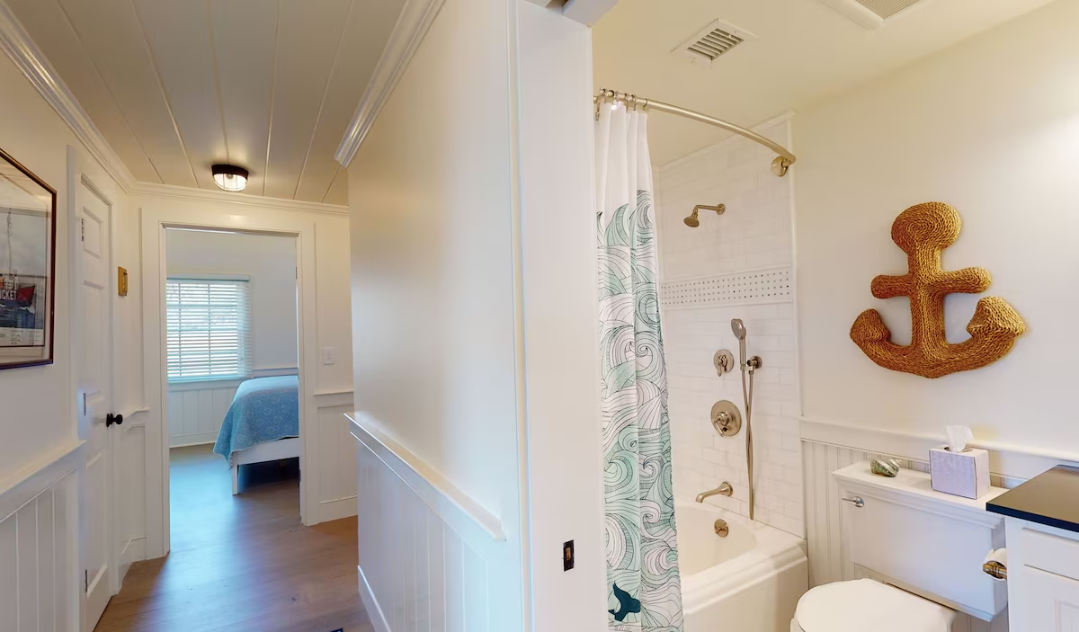 A bathroom off the beadboard hallway with a subway-tiled tub and shower behind a curved brass rail and a green wave-print curtain, a large rope anchor on the wall, and a bedroom visible through the open door
