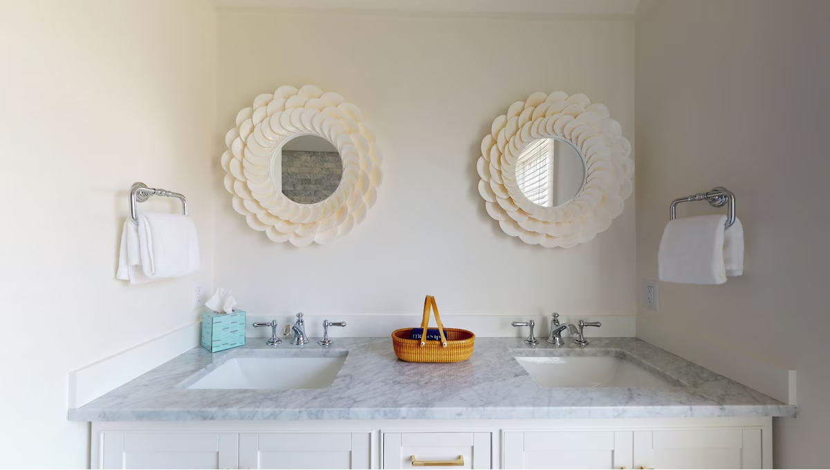 The master vanity head-on — two shell-petal mirrors over a marble counter with chrome cross-handle faucets, towel rings either side, and a Nantucket basket set between the sinks