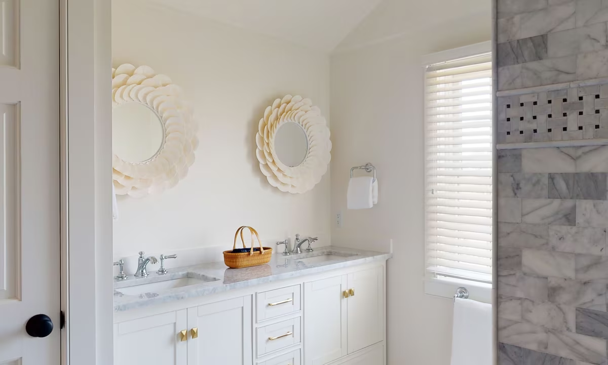 The master bathroom: a white shaker double vanity with a marble top and brass pulls, two round capiz shell mirrors above it, and a marble-tiled shower with a basketweave accent band beside a shuttered window