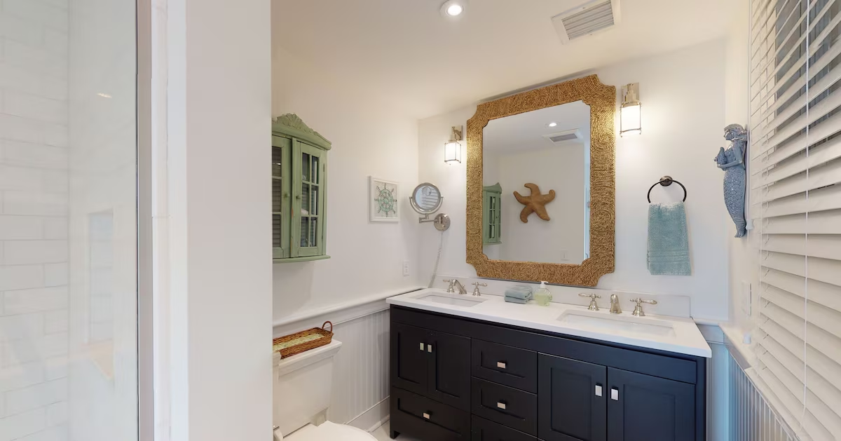 A second bathroom with a navy double vanity under a thick rope-framed mirror, a sisal starfish on the wall, a green painted wall cabinet, a mermaid wall figure, and a white subway-tiled shower