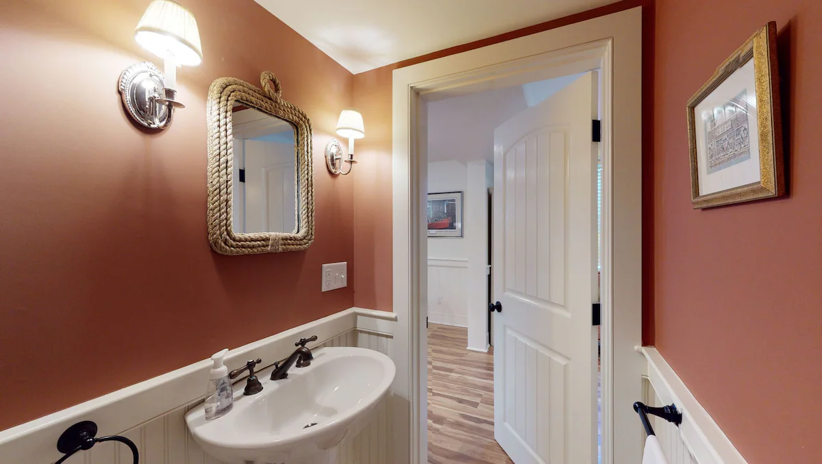A narrow powder room with terracotta walls above cream beadboard wainscot, a pedestal sink with an oil-rubbed bronze faucet, a thick rope-framed mirror between two chrome sconces with pleated shades, and an open door to the hall