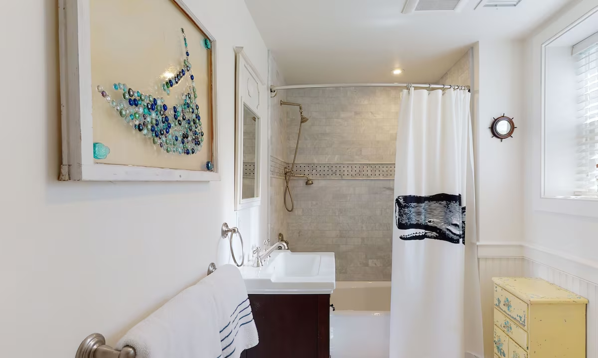 A long white bathroom with a marble subway tub-and-shower behind a curved rail and a whale-print curtain, a mosaic band running through the tile, a sea-glass whale mosaic in a weathered frame on the near wall, a ship’s-wheel porthole mirror by the window, a dark vanity with a white sink, and a distressed yellow chest at the end