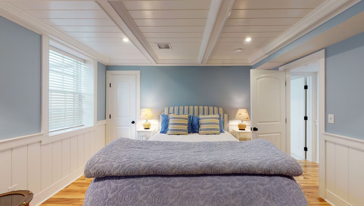A blue-walled bedroom under a coffered beadboard ceiling, with a blue-and-yellow striped upholstered headboard, matching shams, a lavender matelassé quilt, twin gourd lamps on white nightstands, and beadboard wainscoting below