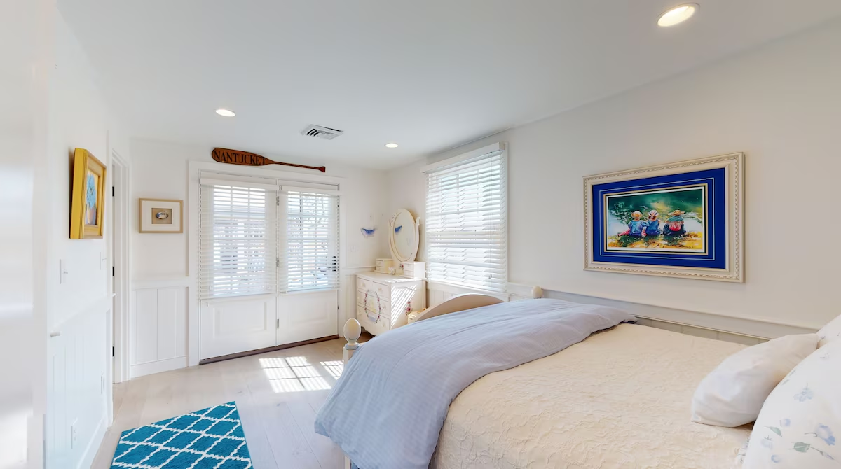 A bright bedroom with a pale blue duvet over a cream quilted bed, shuttered French doors to the garden, a NANTUCKET oar mounted above them, a white dresser with an oval mirror, and a turquoise trellis rug on whitewashed floors