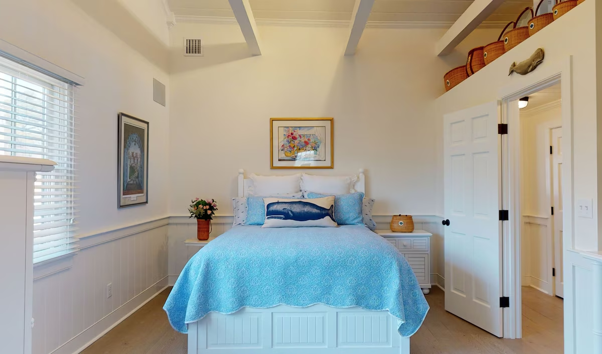 A guest bedroom with a white cottage bed under a pale blue quilt and whale pillow, beadboard wainscoting, framed floral watercolors, and lightship baskets lined up above the doorway