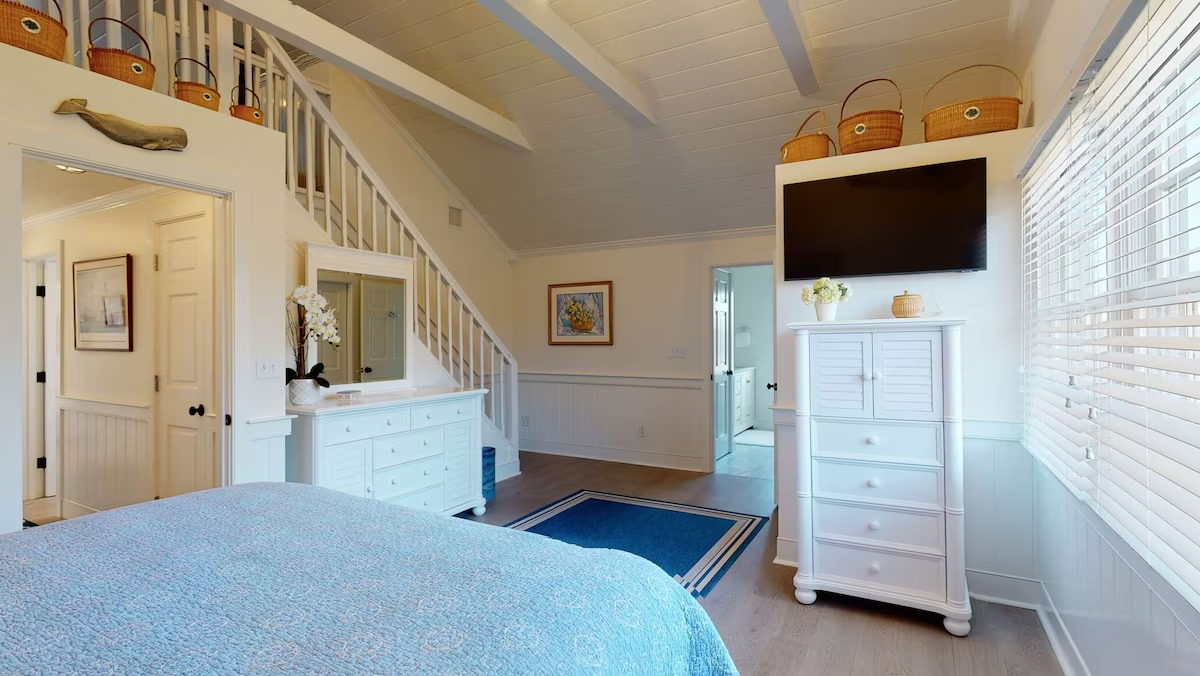 The same loft bedroom from the other side: a carved whale plaque over the doorway, lightship baskets along both ledges and the stair rail, a white dresser with mirror, a tall louvered chest under a wall-mounted television, and a blue rug on the plank floor