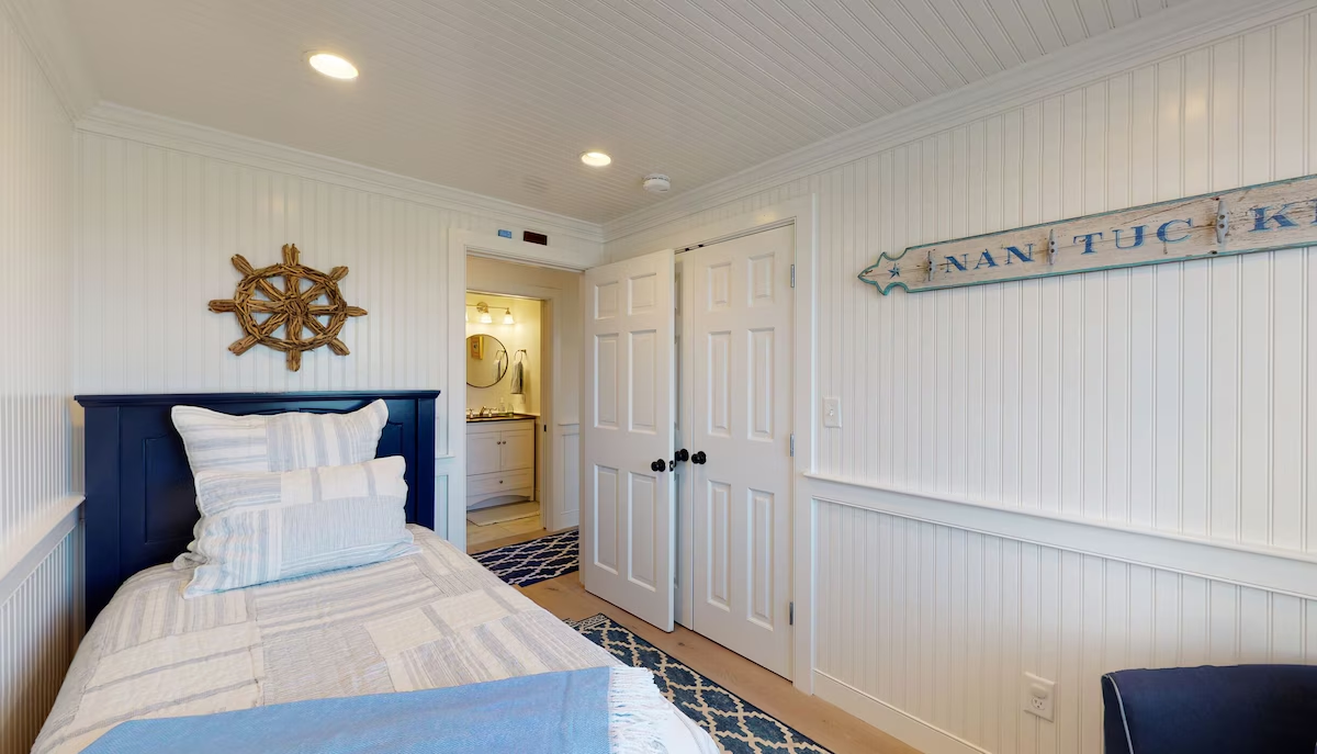 A navy panel bed against full white beadboard walls, with a rope ship’s wheel above it, a long weathered NANTUCKET plank sign on the opposite wall, a double closet, and an ensuite bathroom vanity through the open door