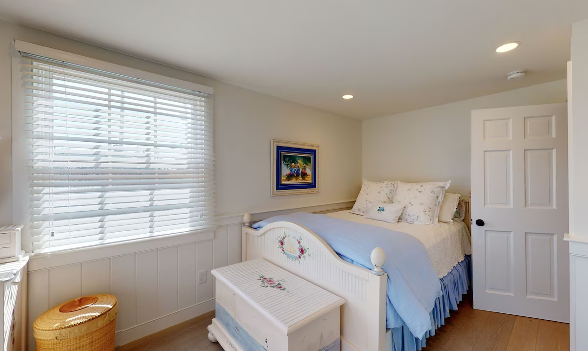 A small bedroom with a hand-painted white cottage bed — a sailboat on the footboard and roses on the blanket chest — made up in blue with a ruffled skirt and floral pillows beneath a wide shuttered window