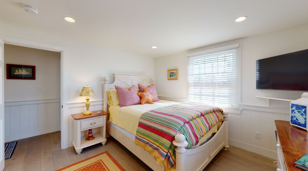A white bedroom with an X-detail cottage bed, a rainbow striped throw folded across the foot, pink chevron and starfish pillows, a wall-mounted television, and a ship print in the hall through the open doorway