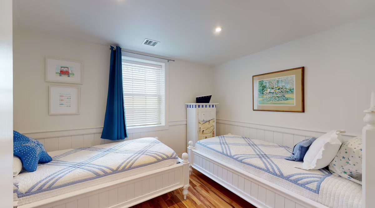 A twin room with two white cottage beds made up in blue plaid quilts, beadboard wainscoting, a blue curtain at the shuttered window, a hand-painted beach-scene chest between the beds, and framed prints on the wall