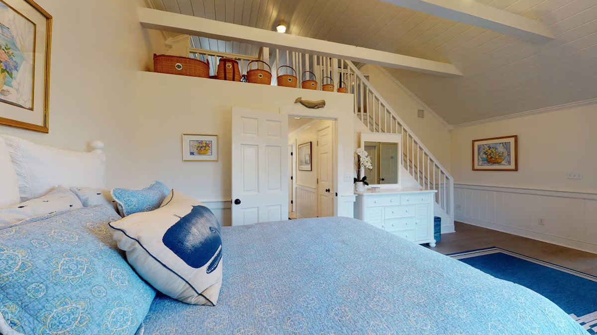 A bedroom under a beamed plank ceiling with a blue quilted king bed, a whale pillow, a white staircase rising to a loft, a row of Nantucket lightship baskets on the ledge above, and a white dresser