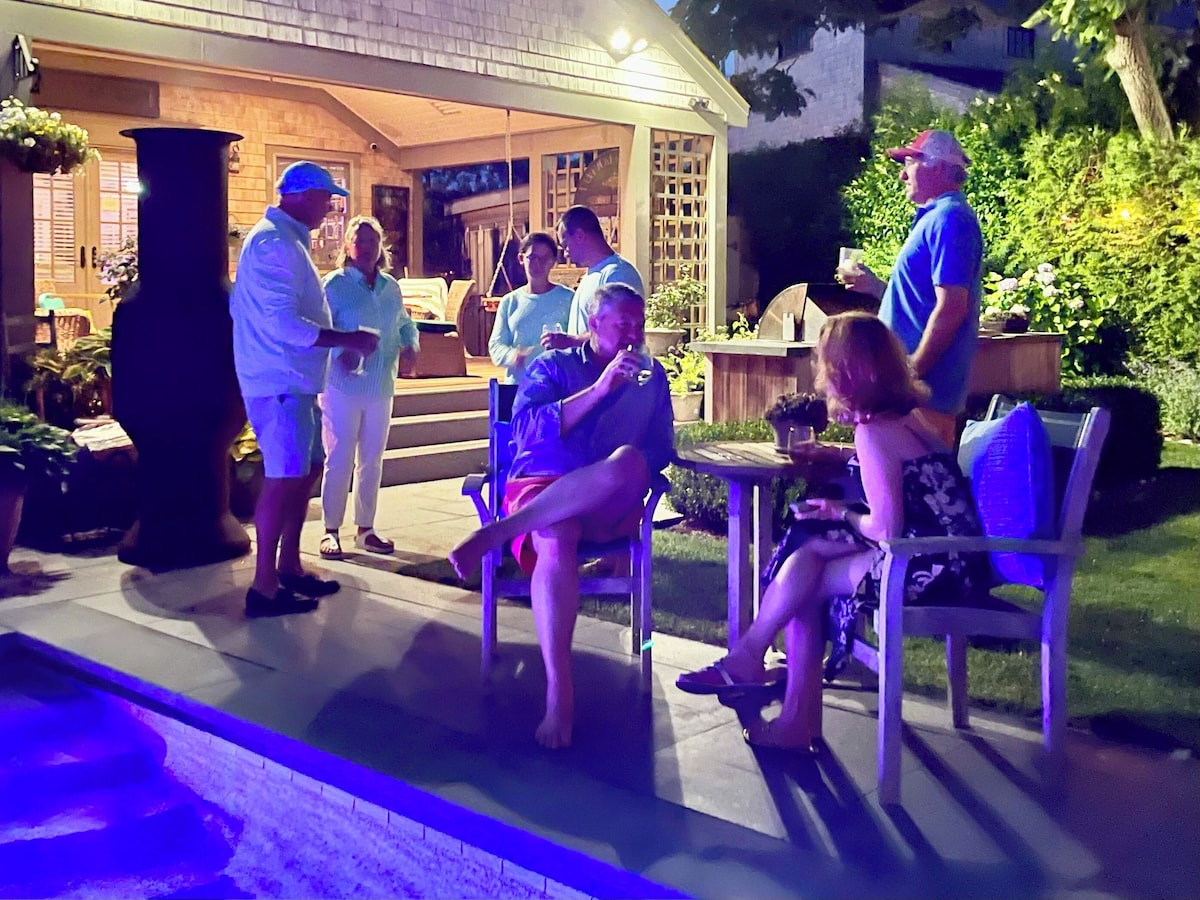 The same evening from the other end: guests standing and sitting with drinks on the bluestone by the lit pool, the tall black chiminea alight, and the shingled pool house glowing warm behind them with its hanging swing and bar counter