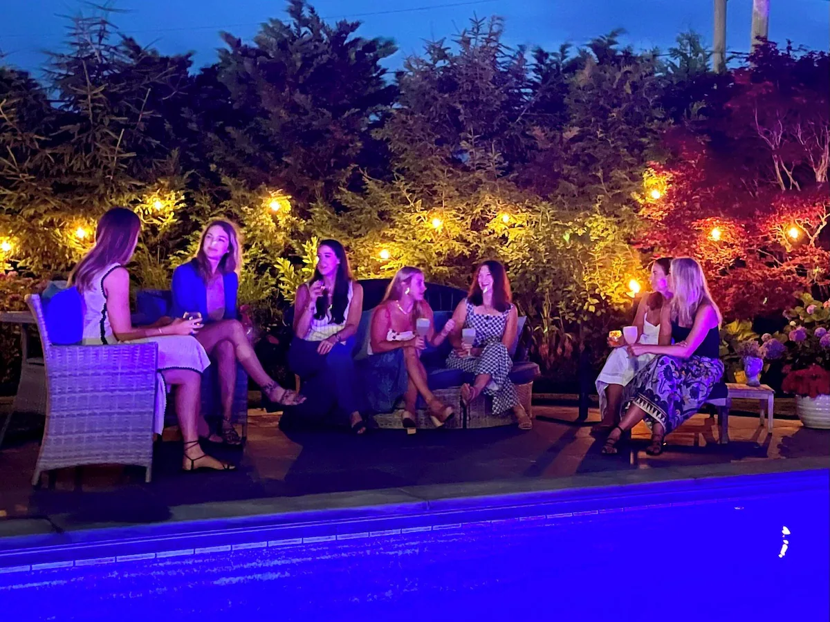 The garden after dark: guests sitting with drinks on wicker sofas and armchairs along the pool patio, string lights and uplit evergreens and a red maple behind them, and the lit blue water of the pool in the foreground