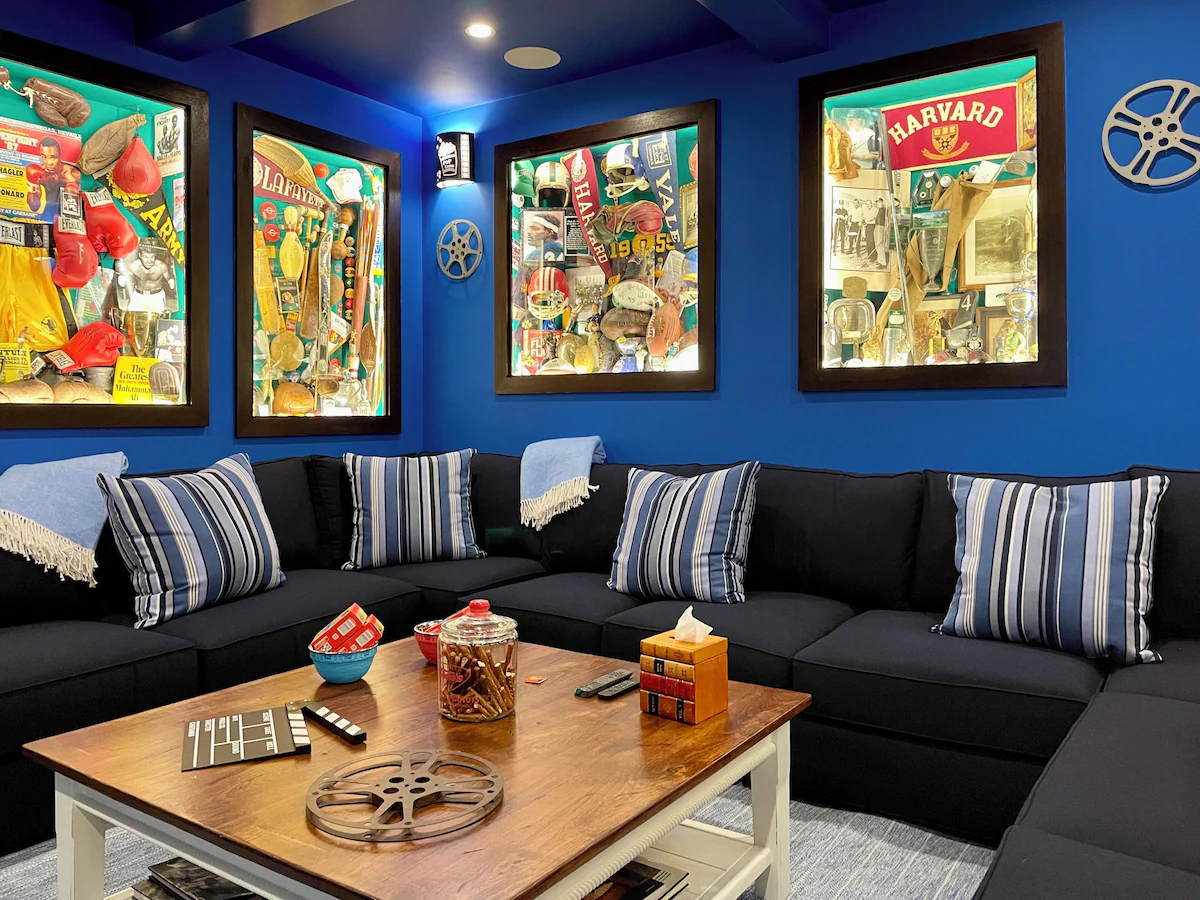 Four of the lit shadow boxes head-on — boxing gloves and Ali, Lafayette oars and medals, Harvard and Yale helmets and pennants — over the sectional, with a clapperboard, candy bowls and a pretzel jar on the wooden coffee table