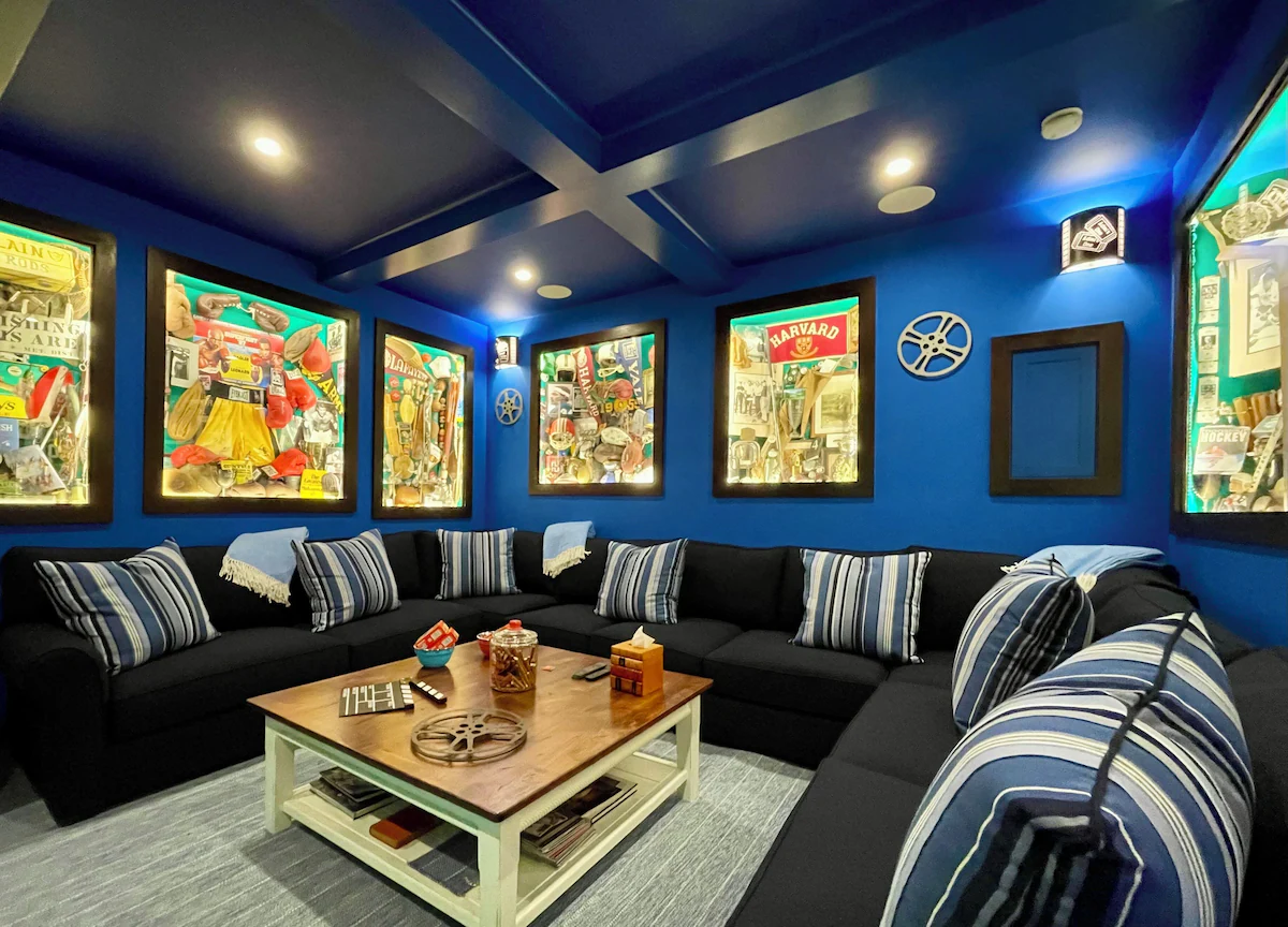 The movie room from the far corner, showing the coffered blue ceiling, the full wraparound of the sectional, a film-camera sconce lighting the wall, and the cases running down both sides of the room