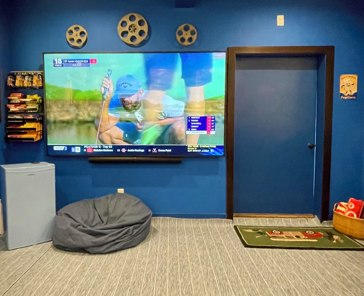 The screen end of the movie room: a large flat screen showing golf under three mounted film reels, a wall rack stocked with candy, a mini fridge, a gray bean bag on the carpet, and a golf-themed toss rug with a basket of red bags beside the door