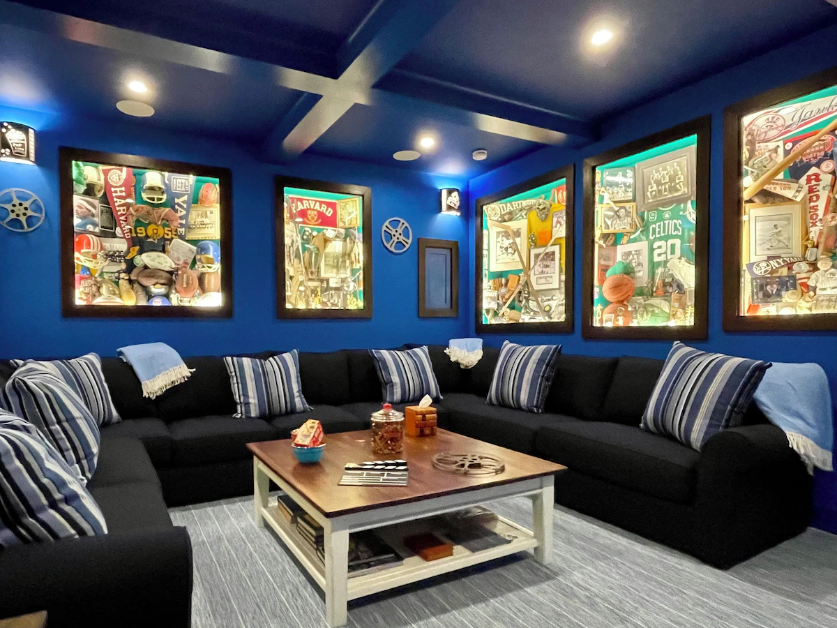 The movie room from the doorway: a black sectional wrapping three walls with blue striped pillows and pale blue fringed throws, cobalt walls hung with six backlit memorabilia shadow boxes — Harvard, Yale, Dartmouth, Celtics, Red Sox, Yankees — cast film reels between them, a coffered blue ceiling with recessed lights, and a white farm coffee table on a gray striped rug