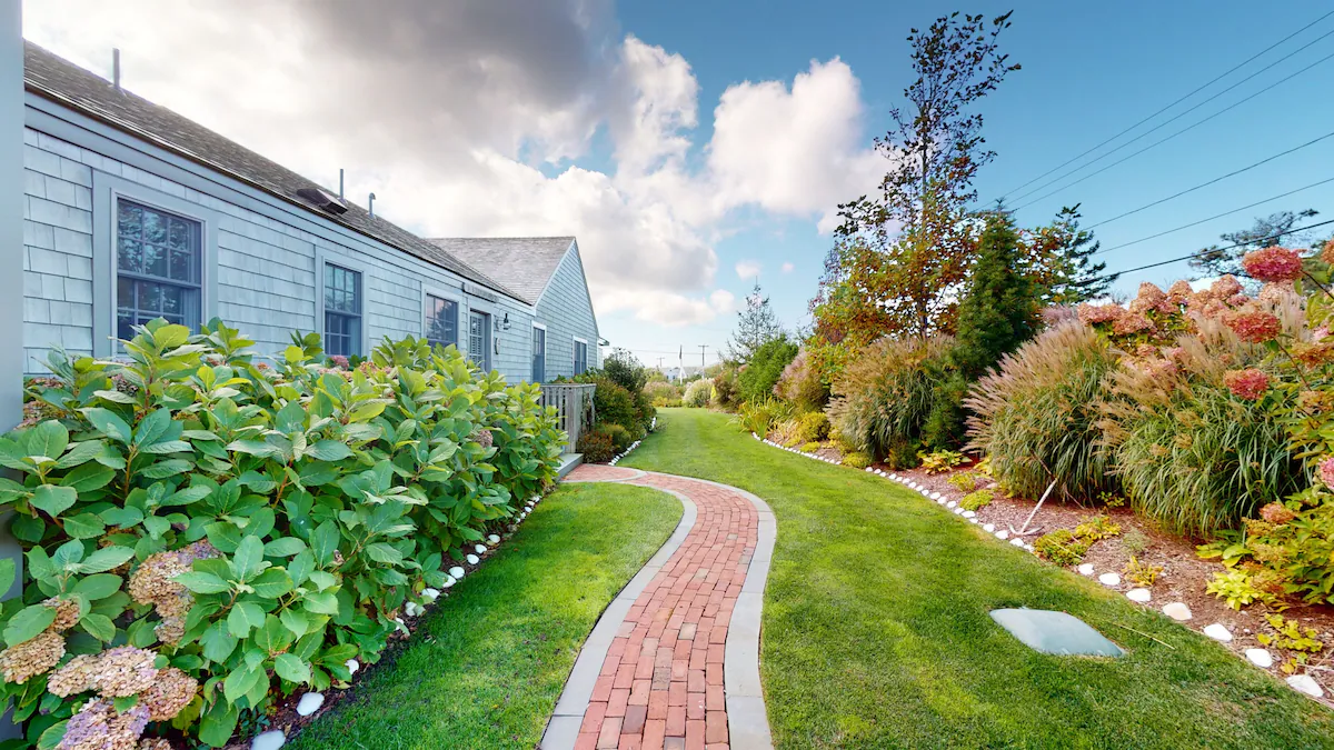 A brick paver path curving between the pale blue-gray shingled side of one of the buildings and a bed of ornamental grasses, with a long hydrangea border along the wall and mown lawn on both sides