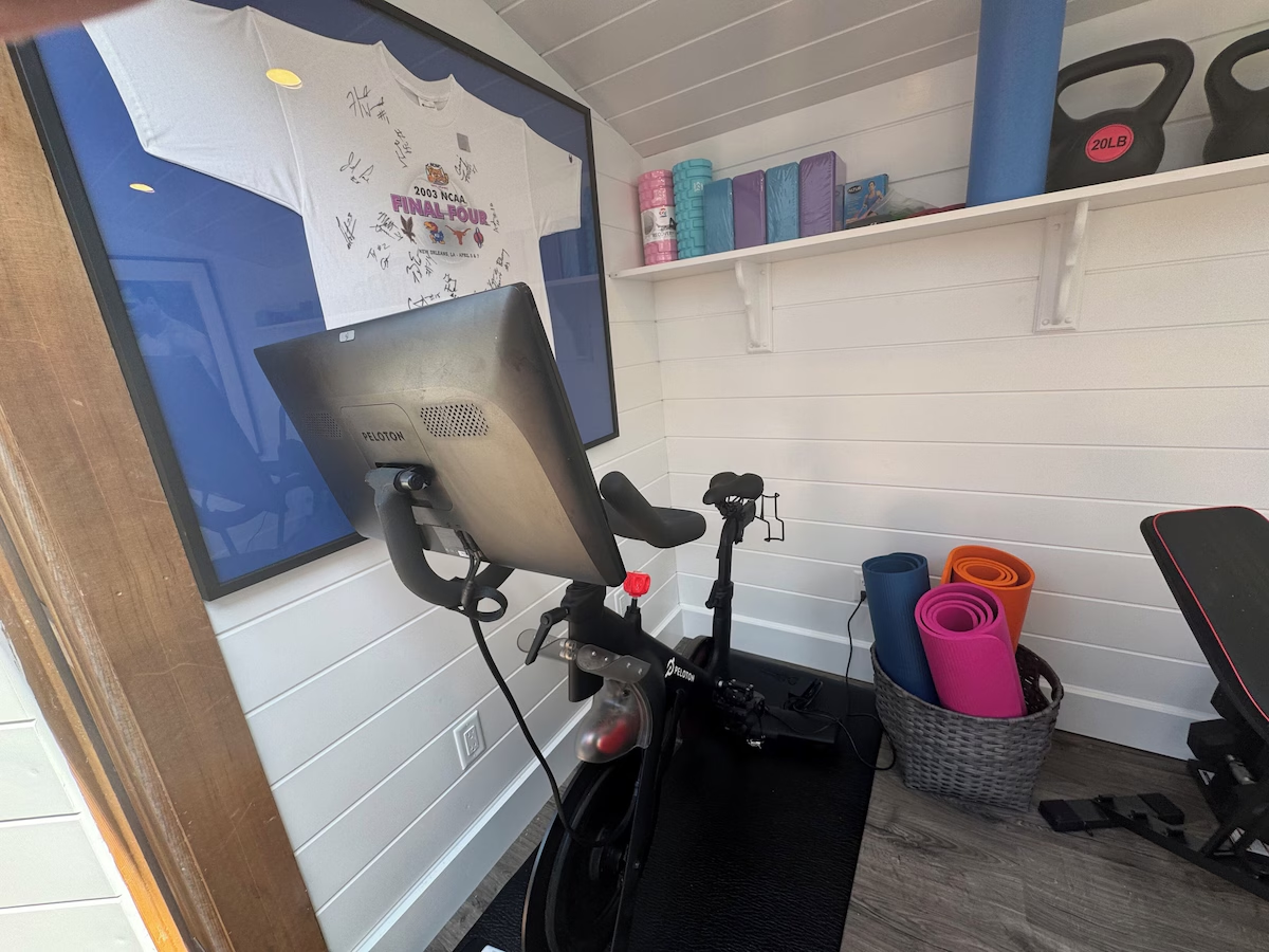 The Peloton bike facing a framed 2003 NCAA Final Four jersey covered in signatures, with kettlebells and stacked yoga blocks on the shelf above and rolled mats in a basket beside it