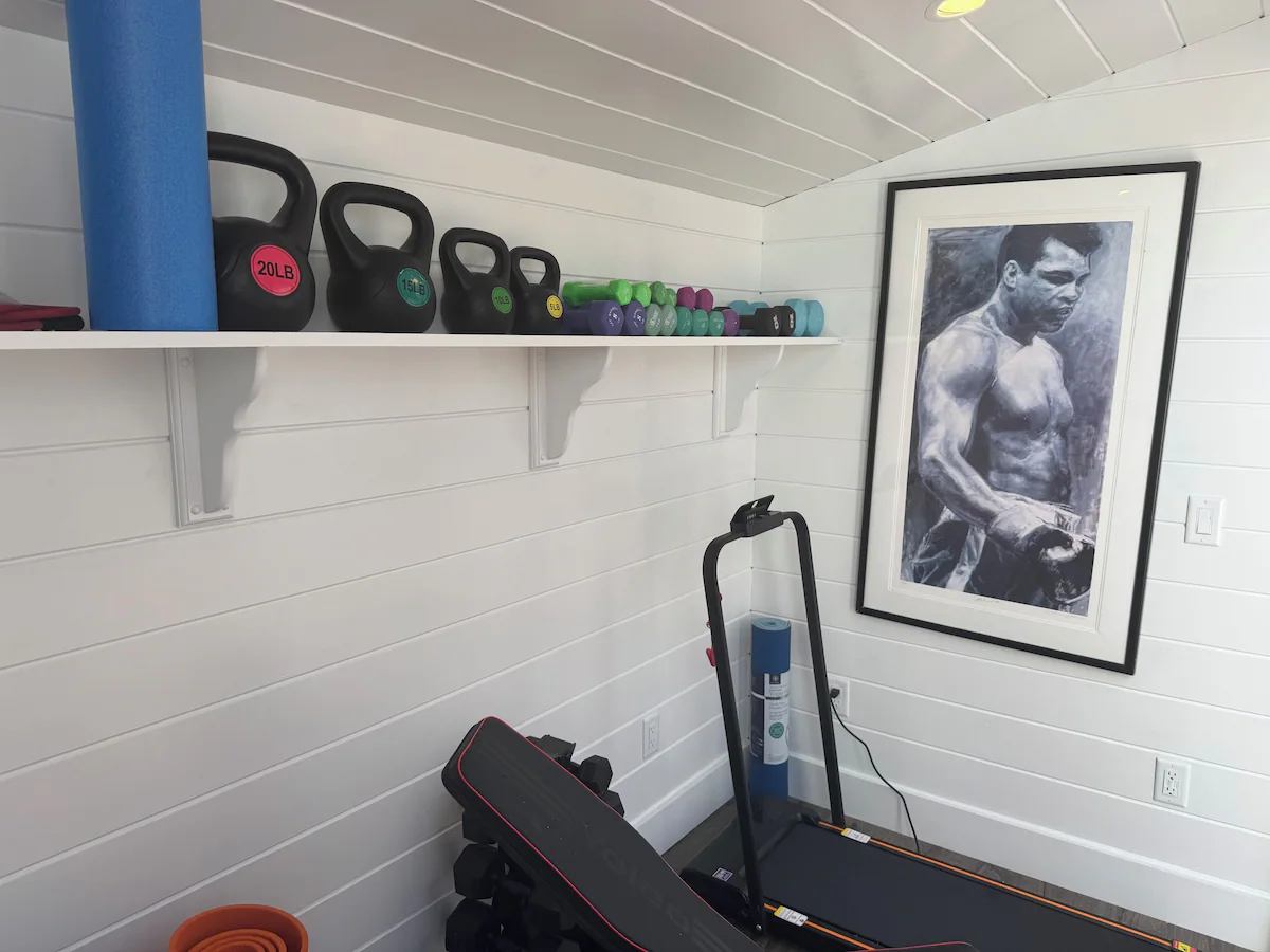 The other end of the gym: kettlebells stepping down from 20 to 5 pounds beside a row of colored dumbbells on a white shelf, a folding treadmill and weight bench below, and a framed black-and-white painting of Muhammad Ali on the far wall