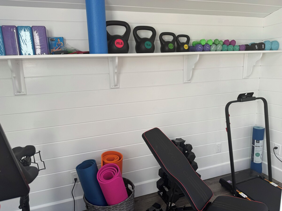 The gym: a long white shiplap wall under a vaulted ceiling with a shelf of kettlebells and neoprene dumbbells above, an adjustable bench and dumbbell rack, a folding treadmill in the corner, a Peloton bike at the edge of frame, and rolled yoga mats in a basket on the floor