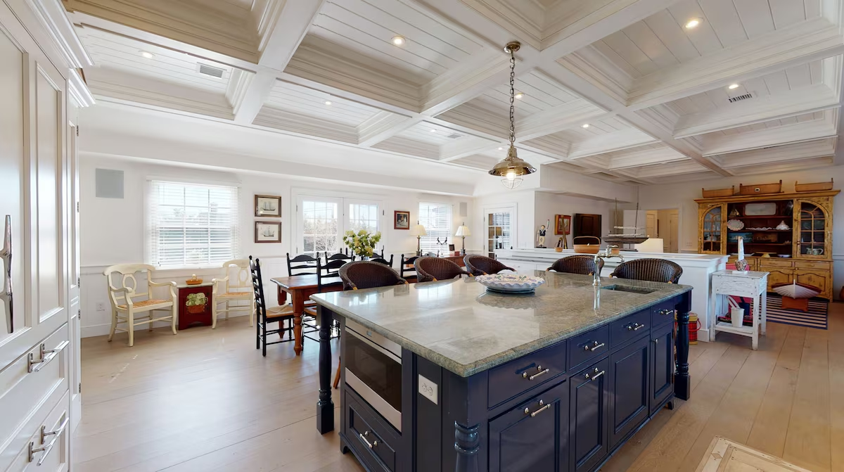 The kitchen open to the dining table and its black ladderback chairs, with framed ship prints, a pair of painted armchairs by the window, and the antique hutch beyond